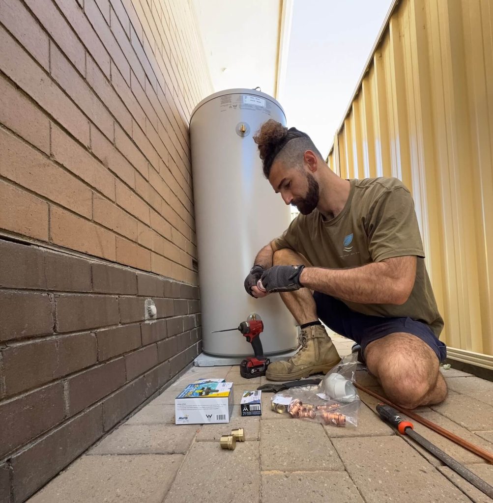 Taro Flow Plumber Matt Taro inspecting a large hot water storage cylinder mounted against a brick wall in an outdoor passage